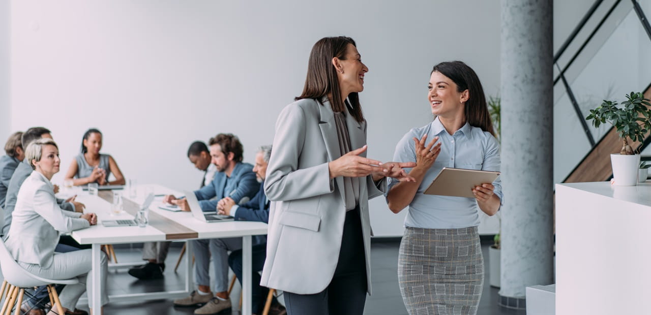 Two colleagues talk while holding a tablet in a modern conference room, with a group of people seated and working at a table in the background.