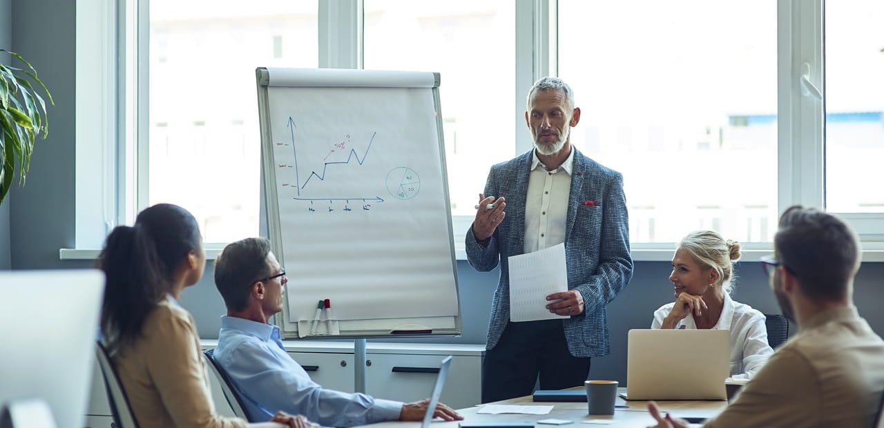 A business owner leads a meeting in a conference room, speaking to colleagues seated around a table with a flip chart showing a line graph.